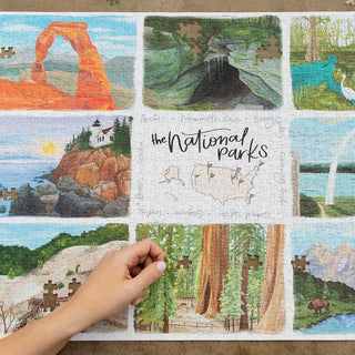 A hand places a puzzle piece into a partially completed jigsaw puzzle titled The National Parks, featuring illustrations of various national park scenes.