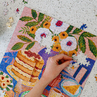 A hand placing a piece into a partially completed jigsaw puzzle featuring a painted illustration of a layered cake and flowers.