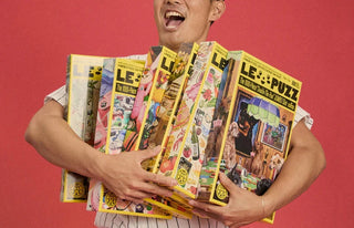A woman sitting on a beige sofa smiles and holds up a puzzle box while holding two more puzzle boxes on her lap.