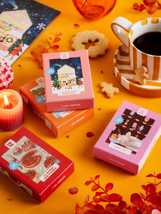 Four small puzzle boxes with illustrated covers are arranged on an orange table with a striped cup of coffee, cookies, a candle, and decorative red foliage.