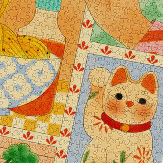 A close-up of a jigsaw puzzle featuring a drawing of a waving white cat with a red collar and other colorful objects and patterns in the background.