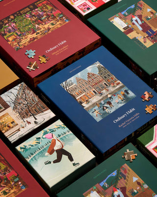 Several colorful jigsaw puzzle boxes with illustrated winter and holiday scenes are arranged closely together, with loose puzzle pieces placed on top of some boxes.