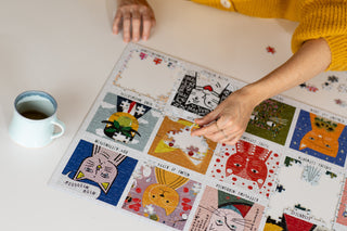 A person in a yellow sweater assembles a colorful jigsaw puzzle featuring illustrated animal faces on a white table next to a light blue mug with a drink.