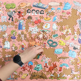 A person with a black watch places a piece in a colorful, partially completed jigsaw puzzle featuring various cartoon food and character illustrations on a pink and cork background.