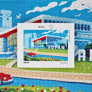 A completed jigsaw puzzle showing a modern building labeled ROYAN, with a car on a road, flowers, and a blue sky, alongside the puzzle box featuring the same image.