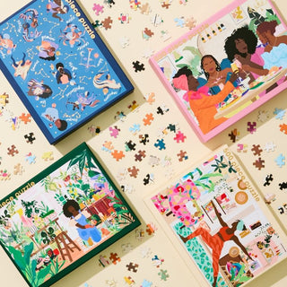 Four colorful jigsaw puzzle boxes with vibrant illustrations are displayed on a surface scattered with loose puzzle pieces.