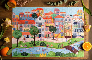 A completed jigsaw puzzle showing a colorful illustration of buildings, trees, and people by a riverside, surrounded by flowers and orange slices on a wooden table.