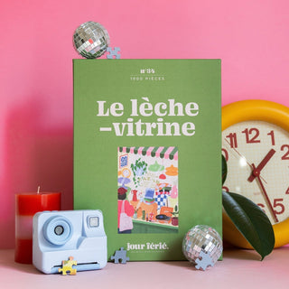 The Jour Férié Le Lèche-Vitrines 1,000 piece jigsaw puzzle box features a shop window illustration surrounded by a disco ball, a yellow clock, a light blue instant camera, a candle, and loose pieces.