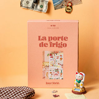 A pink La Porte de Frigo | 1,000 Piece Jigsaw Puzzle by Jour Férié stands upright on a peach background, surrounded by a figurine, fabric, ribbon-cut puzzle pieces, and pinned photos above.
