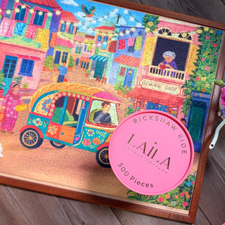 Rickshaw Ride | 500 Piece Jigsaw Puzzle by Laila Luxury Puzzles features a vibrant Pakistani market with a rickshaw and people; its box rests on one corner of the completed, colorful scene.