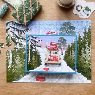 A nearly finished All Packed | 300 Piece Jigsaw Puzzle by ViSSEVASSE depicts a van filled with gifts winding through a snowy forest, with a coordinating holiday card placed on top of the puzzle scene. Surrounded by wrapped presents and crafting supplies, this puzzle makes for the perfect festive activity.