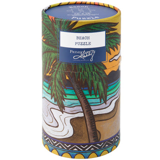 The Primitives by Kathy Beach | 500 Piece Jigsaw Puzzle comes in a cylindrical box with colorful artwork of a palm tree, ocean waves, and a sunset.