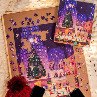 The partially completed Holiday Hotspot 500 Piece Jigsaw Puzzle by Chuffed Puzzles, featuring a city ice rink and Christmas tree with the box displaying the finished image, is set on a cream textured surface.