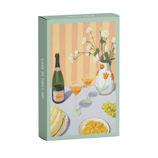 Love At First Sip | 99 Piece Jigsaw Puzzle by Piecely features vibrant textured art by a Lithuanian artist—a pastel recycled cardboard box with an illustration of a table set with sparkling wine, fruit, and flowers.