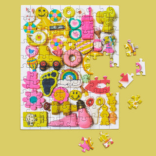 Lemonade | 100 Piece Jigsaw Puzzle by WerkShoppe features Lisa Congdon’s colorful frosted cookies—smiley faces and rainbows—on a yellow background, with recycled board pieces scattered nearby.