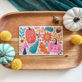 The Spaghetti Tree Happy Fall 150 Piece Jigsaw Puzzle, featuring a vibrant pumpkin design, is completed on a wooden tray and surrounded by decorative pumpkins, acorns, and yellow pom-poms.