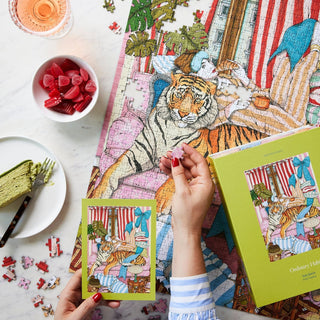 A person assembles the Wild Comfort | 1,000 Piece Jigsaw Puzzle by Ordinary Habit among lush plants, with a drink, green cake, and pink candies nearby.