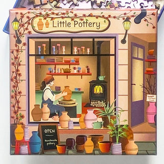 ViSSEVASSE’s “Little Pottery | 150 Piece Jigsaw Puzzle” features a charming pottery shop named Little Pottery, complete with shelves of handmade ceramics, an artist shaping clay at the wheel, lush plants outside, and an OPEN chalkboard sign by the sidewalk—ideal for anyone seeking inspiration or wishing to assemble their own unique scene piece by piece.