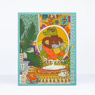 A vibrant box features an alligator in a sweater reading a book in a warmly decorated room. Text reads Rainy Day | 1,000 Piece Jigsaw Puzzle by Birdie Puzzles. Crafted from recycled materials, this puzzle offers an eco-friendly experience.
