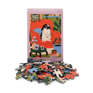 The Happily Jigsaw Puzzles Rabbit 99 Piece Mini Jigsaw Puzzle box features a dog on an orange chair, is made from recycled board, and has loose pieces scattered in front.