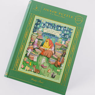 The Good Juju Ink "Snaily's Books | 1,000 Piece Jigsaw Puzzle" comes in a green keepsake box featuring an illustration of a snail reading among piles of books, teacups, and animals, beautifully accented with elegant gold foil details. The box reads: "Snails Slow Living Books & Tea, 1000 pieces.
