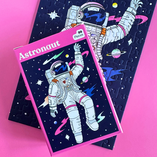 A matte finish Puzzledly Astronaut | 99 Piece Jigsaw Puzzle box resting on a partially assembled pocket-sized puzzle of an astronaut floating in space.
