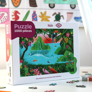 The Pitisdessins | 1,000 Piece Jigsaw Puzzle by Lundi en Huit features an original illustration of people swimming and relaxing near a waterfall in a lush landscape, with the box on a table and puzzle pieces scattered around.