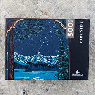Enjoy cozy nights in with the Fireside 500 Piece Jigsaw Puzzle by The Montana Scene, featuring an illustrated nighttime mountain landscape with pine trees, stars, and a lake—a perfect Montana-themed puzzle.