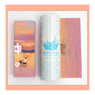 A Momibook Sunset Artist 1,000 Piece Jigsaw Puzzle box, a rolled puzzle mat, and the finished sunset scene with two people by the water are neatly displayed on a white surface with an orange border.