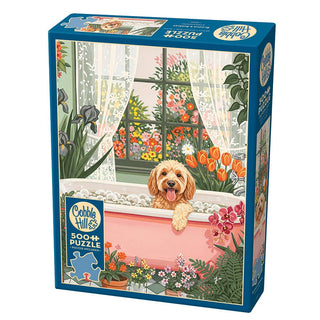 The Cobble Hill "Blooms and Bubbles" 500 piece jigsaw puzzle features a dog in a pink bathtub, surrounded by colorful flowers and a large window—ideal for dog lovers.
