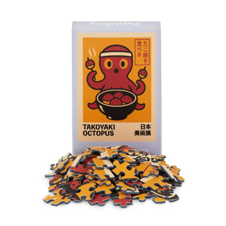 A box labeled "Takoyaki Octopus | 99 Piece Mini Jigsaw Puzzle" by Happily Jigsaw Puzzles, featuring an octopus illustration, sits behind a pile of loose, recycled board puzzle pieces.