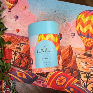 A cylindrical Laila Luxury Puzzles box is placed atop a completed Cappadocia | 500 Piece Jigsaw Puzzle, with green leaves on the left, enhancing the vibrant hot air balloon scene.