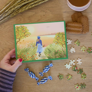A hand holds a ViSSEVASSE postcard featuring nature; nearby on the wooden table are wrapped candies, biscuits, a drink, cereal stalks, and pieces from the Autumn | 300 Piece Jigsaw Puzzle.