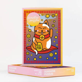The Lucky Cat | 500 Piece Jigsaw Puzzle by Sunshine Puzzles comes in a pastel-colored box featuring a charming cartoon Chinese lucky cat on a red mat, surrounded by stars and coins—making it the ideal puzzle for cat enthusiasts.