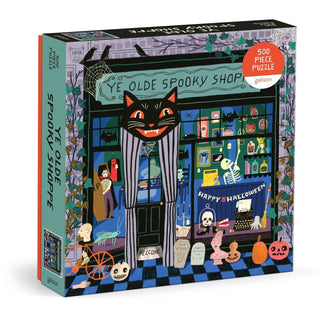 The Galison "Ye Olde Spooky Shoppe" 500 Piece Jigsaw Puzzle features a charming folk-art style illustration of a Halloween-themed shop, adorned with spooky decorations, a black cat sign, skeletons, ghosts, and pumpkins.