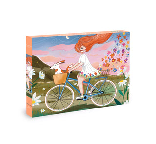 Olleo Puzzles' Blooming Ride is a 500 piece jigsaw puzzle featuring a girl with long red hair riding a blue bike with her dog in the basket, set amid blooming hills and flowers beneath a pink sky.
