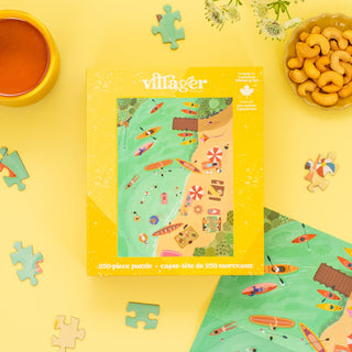 A Lake Day | 250 Piece Jigsaw Puzzle by Villager Puzzles with a beach scene rests on a yellow table among loose pieces, cashews, a drink, and flowers—making it an ideal Canadian souvenir.