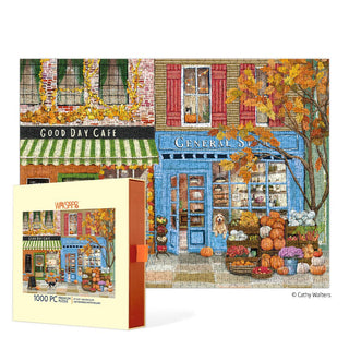The WerkShoppe "Autumn On Main" 1,000-piece jigsaw puzzle features a fall street scene with a cafe, general store, vibrant autumn trees, pumpkins, and a dog in the shop window. The puzzle box appears prominently in the foreground.