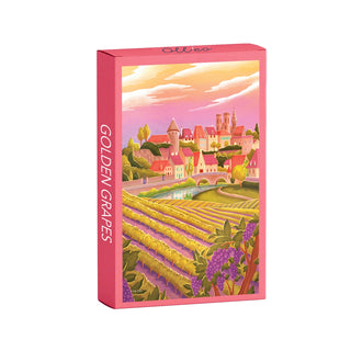 A box labeled Olleo Puzzles Golden Grapes | 99 Piece Jigsaw Puzzle features an illustrated French vineyard scene with grapevines and vibrant village buildings under a warm sky.