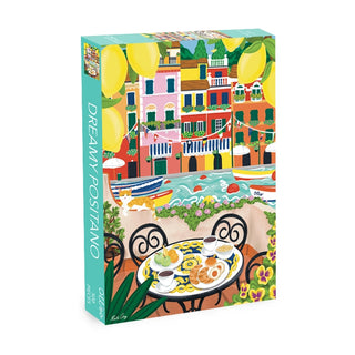The Dreamy Positano 500 Piece Jigsaw Puzzle by Olleo Puzzles features a vibrant seaside scene with boats, bright buildings, lemons, and a table set with pastries, fruit, and coffee.
