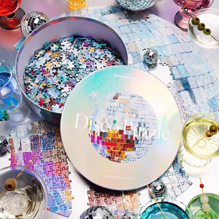 A round box labeled "Disco | 750 Piece Jigsaw Puzzle" by Piecework Puzzles sits open, its holographic pieces spilling onto a partially completed puzzle and a table with cocktails and olives.