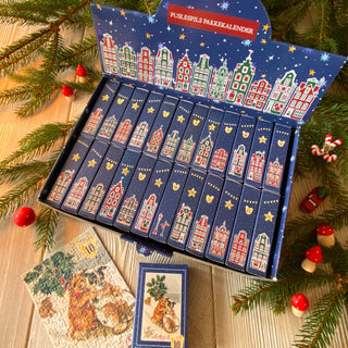 The Penny Puzzle Vintage Postcards Advent Calendar features 24 illustrated drawers, each holding a 99-piece jigsaw puzzle for festive Holiday Countdown fun on a wooden surface with pine branches and Christmas decorations.