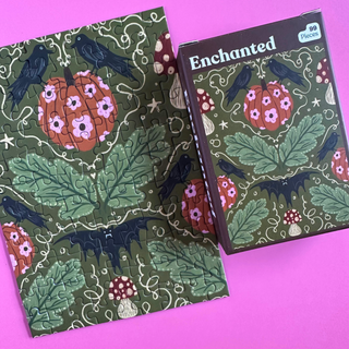 Enchanted | 99 Piece Jigsaw Puzzle, a pocket-sized 99-piece puzzle and its box, featuring an illustration of crows, a bat, floral-patterned pumpkins, and mushrooms on a green background.