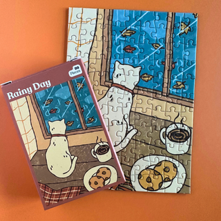 A Puzzledly Rainy Day 99 Piece Jigsaw Puzzle shown with its mini box, featuring a cat looking out a window at a rainy day, with a coffee cup and cookies on a table.