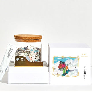 The Skiers is a 450-piece jigsaw puzzle by Jiggy Puzzles, featuring Amanda Oleander’s art of two people on a ski lift in snowy mountains. Includes pieces in a cork-top jar, glue tube, metal tool, and illustrated box.