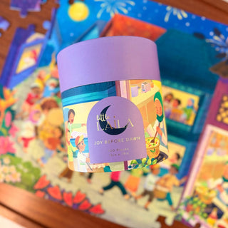 A cylindrical box labeled "Joy Before Dawn | 100 Piece Jigsaw Puzzle" by Laila Luxury Puzzles rests on a partially finished, colorful puzzle themed around family and Indonesia suhoor.