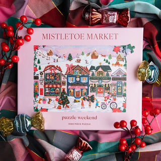 Mistletoe Market 1,000 Piece Jigsaw Puzzle box featuring an illustration of a pink Christmas village with shops and a post office, resting on a plaid fabric background with decorative red berries.
