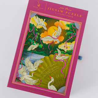 Experience the beauty and sophistication of the Good Juju Ink Birds of A Feather | 500 Piece Jigsaw Puzzle, featuring an enchanting illustration of white cranes soaring around a fan amidst a vibrant sunset and lush green foliage. Housed in a magnetic keepsake box embellished with elegant gold foil accents, this puzzle is as stunning as it is enjoyable to assemble.