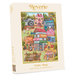 A box of Reverie Puzzles’ Hobby Village 1,000 Piece Jigsaw Puzzle, featuring a charming illustrated village scene with art, knitting, books, gardening, and puzzle shops—a cozy design perfect for fans of relaxing puzzles like the Mushroom Library.