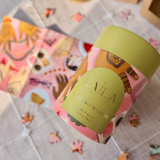 A cylindrical container labeled Laila Luxury Puzzles Sweet Sisterhood | 500 Piece Jigsaw Puzzle sits on a table with a partially completed cultural puzzle and scattered pieces nearby.